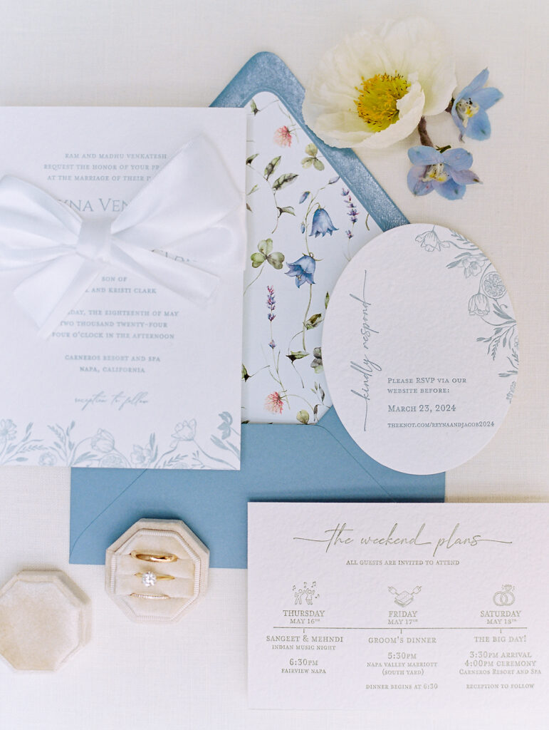 A wedding invitation suite with floral designs, an RSVP card, a wax-sealed envelope, a white bow, a flower, and two rings in a hexagonal box arranged on a white surface.