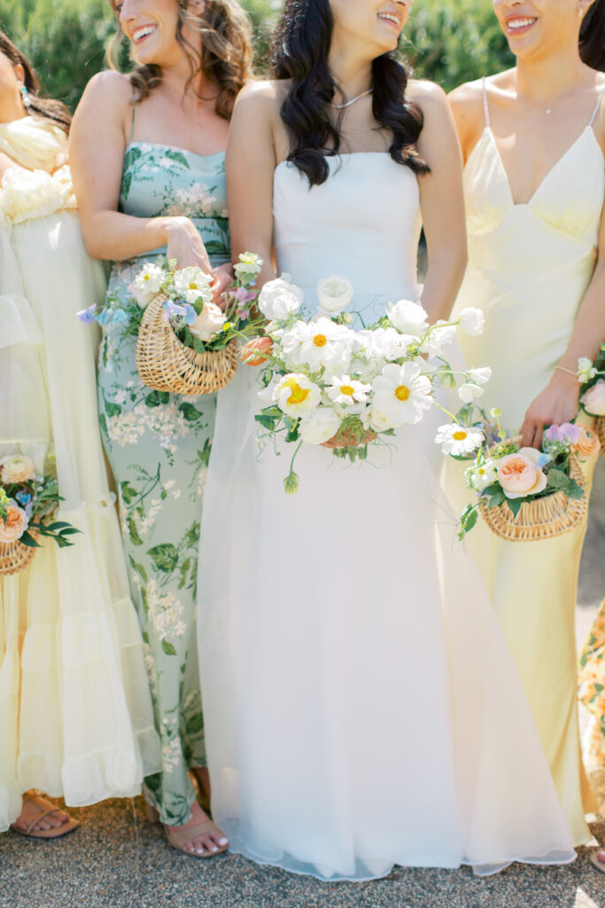 Four women in pastel dresses stand together outdoors, each holding a floral bouquet; the central figure wears a white strapless gown and holds a larger white and yellow flower bouquet.