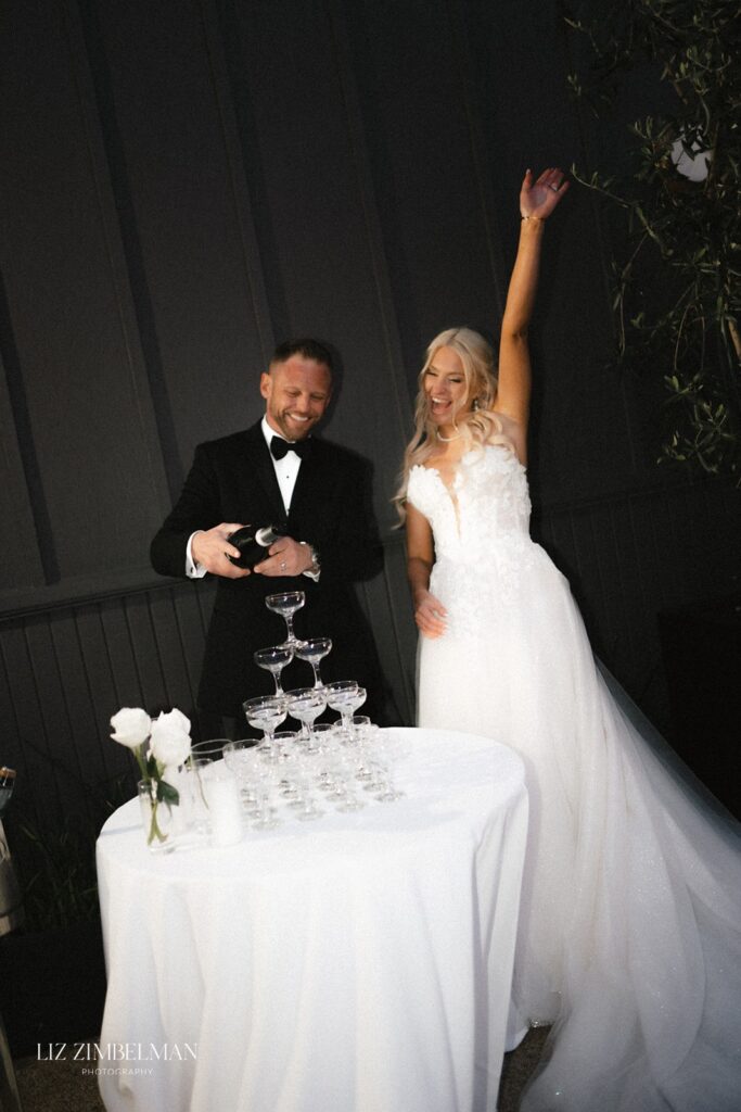 A bride in a white dress raises her arm while a groom in a tuxedo pours champagne into a tower of glasses on a table with white roses.