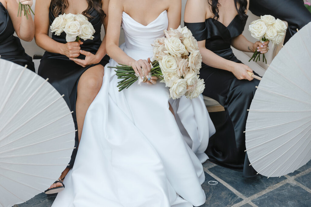 Four women in formal dresses sit with white parasols and hold bouquets of white roses; one woman wears a white gown, while the others wear black dresses.