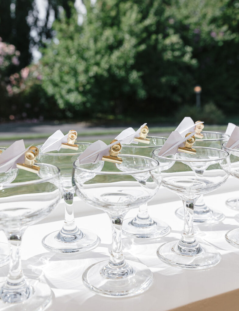 Several empty coupe glasses are arranged in rows, each holding a small white folded card clipped with a gold binder clip, set on a white surface with greenery in the background.