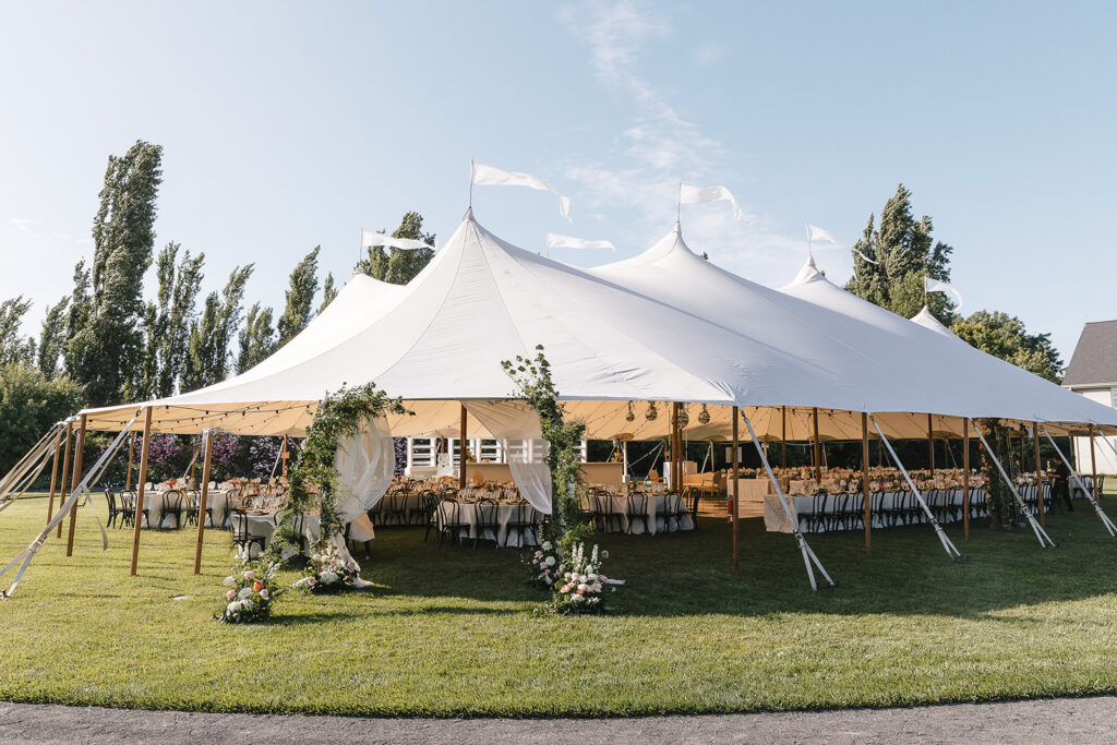 Large white event tent set up on a lawn, decorated with floral arrangements and filled with tables and chairs for a formal outdoor gathering.