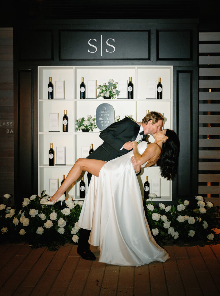 A groom dips and kisses his bride in front of a black and white decorative display with wine bottles and floral arrangements.