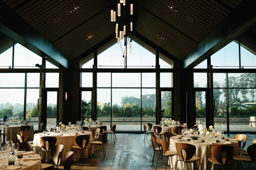 A modern banquet hall with large windows, round tables set for an event, and views of trees outside.