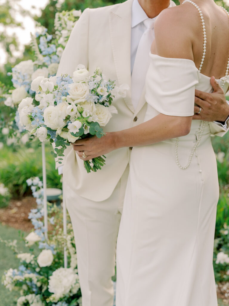A bride in an off-shoulder white dress holding a bouquet of white and blue flowers stands beside a groom in a cream suit and tie.