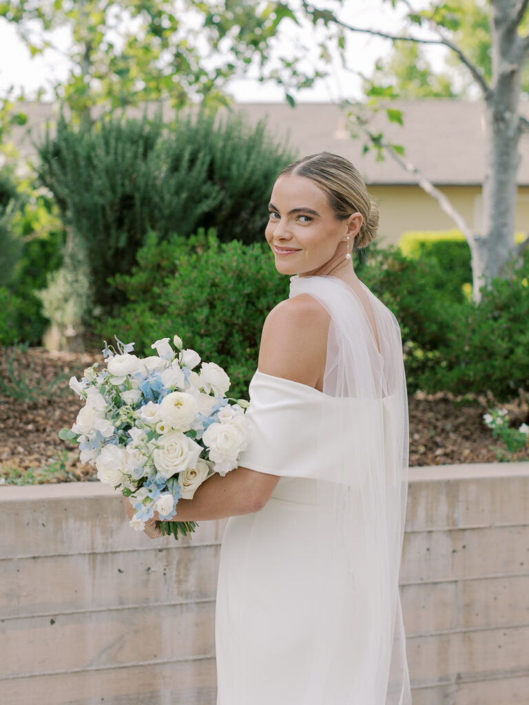 A bride in a white dress holds a bouquet of white and blue flowers, standing outdoors in front of greenery and a low concrete wall.