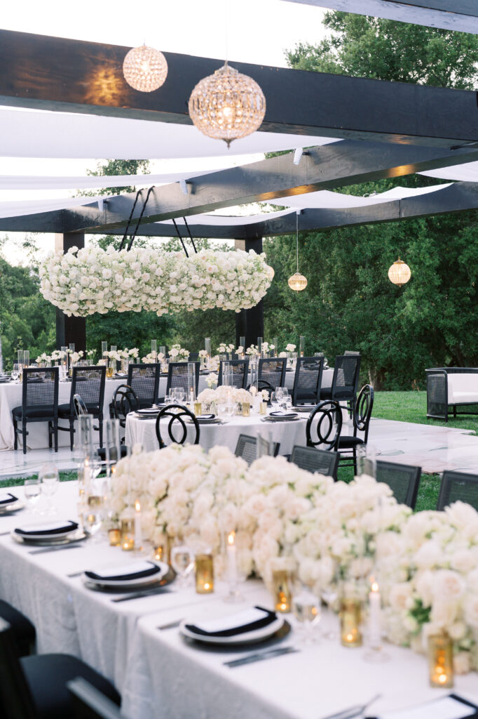 Outdoor wedding reception setup with white floral centerpieces, black chairs, gold candle holders, and hanging chandeliers under a pergola.