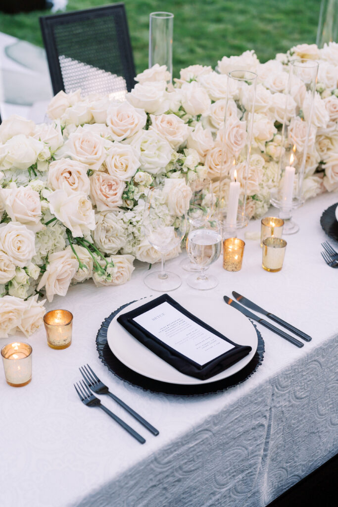 Elegant table setting with white floral centerpiece, black-rimmed plates, black napkins, glassware, gold votive candles, and a printed menu on a white tablecloth.