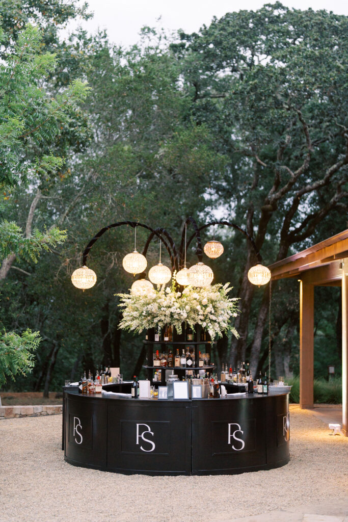Outdoor circular black bar with “FS” initials, decorated with white flowers and hanging globe lights, surrounded by trees and bottles displayed on the counter and shelves.