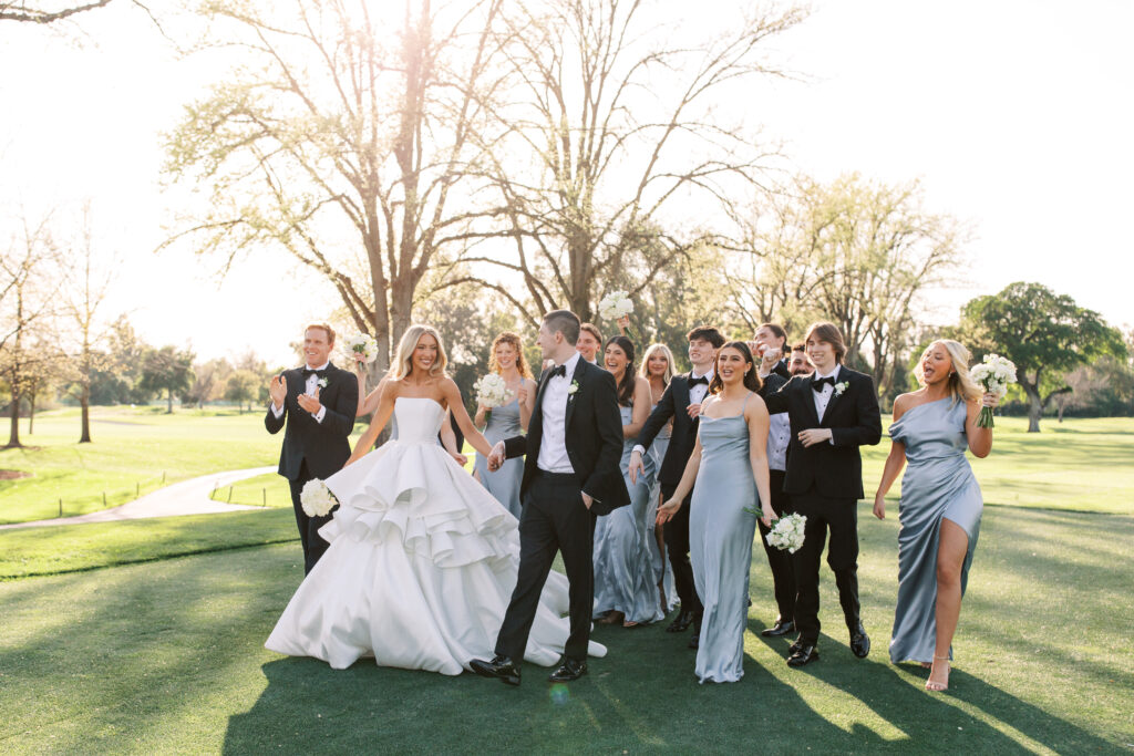 A bride and groom walk with their wedding party outdoors on a sunny day; everyone is dressed formally and holding bouquets.
