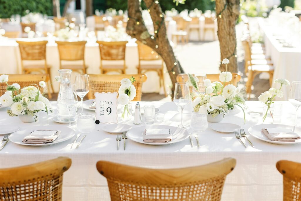 A sunlit outdoor table set for a formal event with white linens, plates, cutlery, glassware, white floral centerpieces, and a table number card reading “9.”.