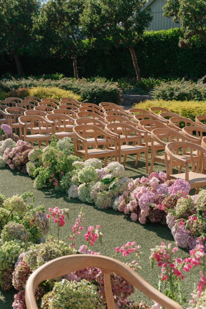Rows of wooden chairs are set up outdoors on grass, surrounded by clusters of pink, white, and green flowers, with trees and shrubs in the background.