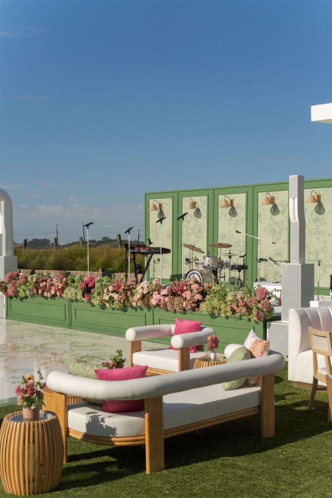Outdoor event stage with green panels, floral arrangements, microphones, and wooden seating featuring white cushions and pink pillows on a grassy area under a clear blue sky.