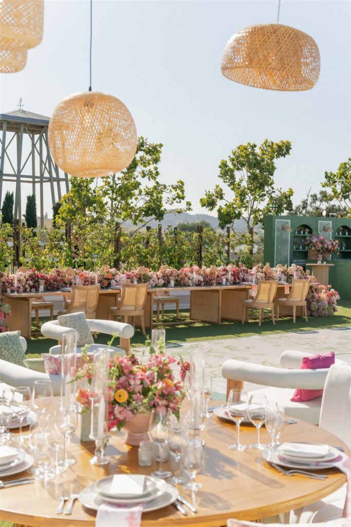 Outdoor event setup with round wooden tables, floral centerpieces, wicker chairs, hanging lights, and greenery in the background under a clear sky.