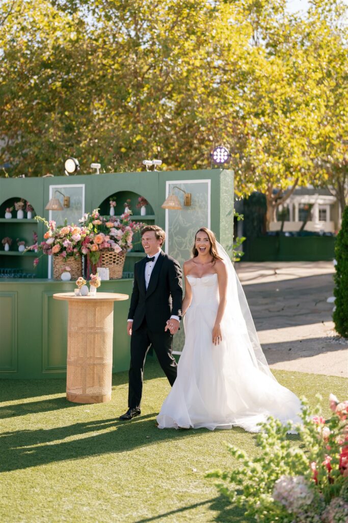 A bride in a white gown and veil walks hand in hand with a groom in a black tuxedo outdoors beside green decor and floral arrangements.