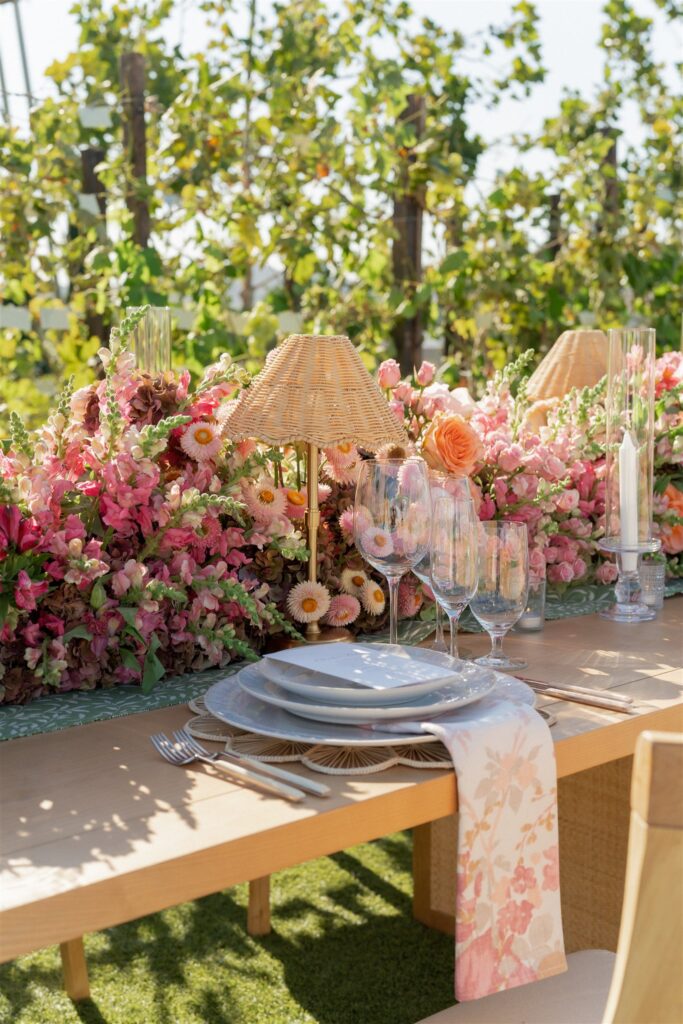 A wooden table set with plates, glassware, floral napkins, and surrounded by pink and peach flowers, small lamps, and outdoor greenery in the background.