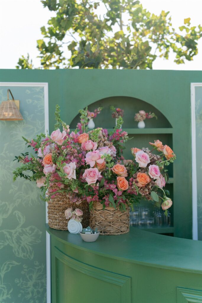 A green counter with woven baskets holding pink and peach roses, with small vases on a green shelf in the background and leafy trees visible outside.