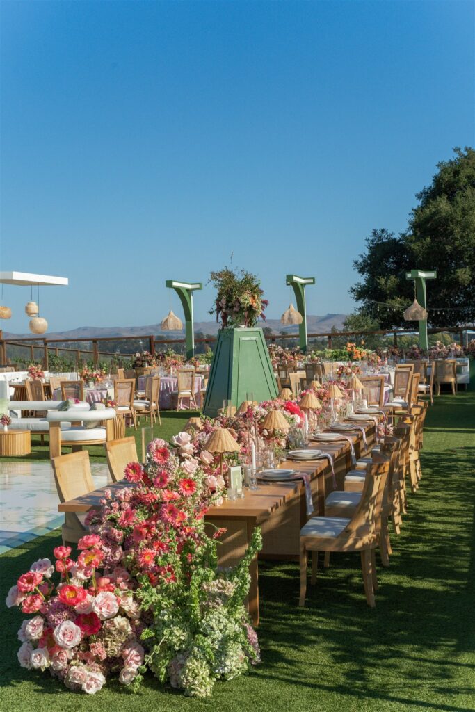 Outdoor event setup with long wooden tables, floral centerpieces, wooden chairs, and surrounding greenery under a clear blue sky.