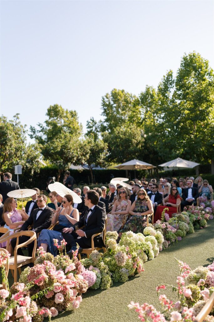 Guests seated outdoors at a formal event, surrounded by flower arrangements, with green trees and umbrellas in the background on a sunny day.