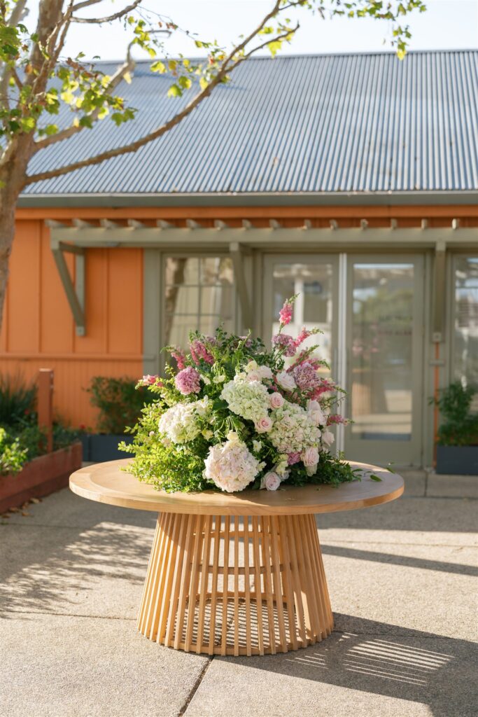 A round wooden table with a large floral arrangement sits outside a building with orange siding and a metal roof on a sunny day.