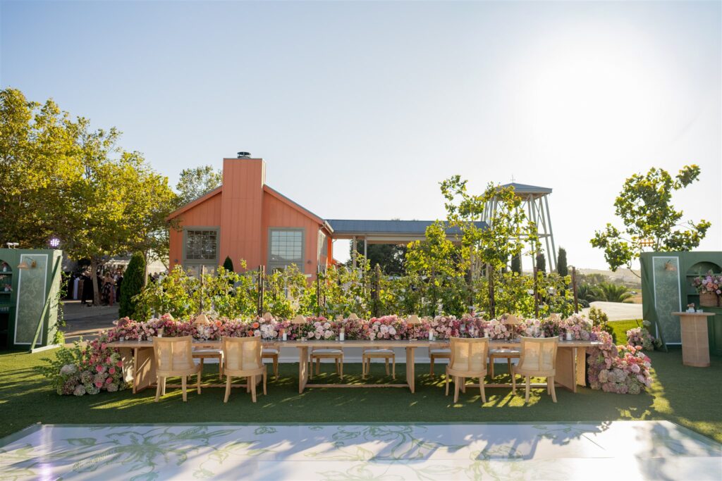 A long outdoor dining table decorated with pink and peach flowers is set on grass in front of a small orange building under a clear sky.