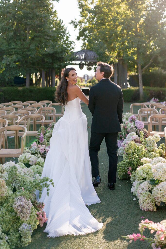 A bride in a white dress and a groom in a black suit walk down an outdoor aisle decorated with flowers and empty chairs on a sunny day.