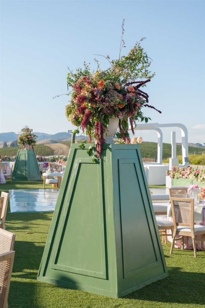 Large green pedestal with a floral arrangement stands outdoors on grass, surrounded by tables and chairs with a scenic landscape and blue sky in the background.