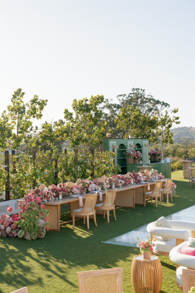 Long banquet table with wicker chairs and pink floral arrangements is set outdoors on grass, with trees and greenery in the background under a clear sky.