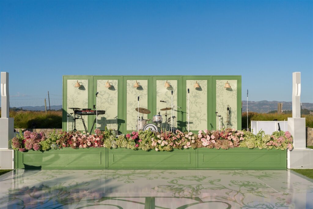 Outdoor stage with green panel backdrop, floral arrangements at the base, musical instruments, and microphones set up, under a clear blue sky.