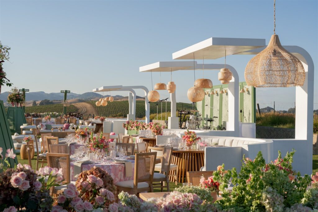 Outdoor event setup with round tables, floral centerpieces, modern white canopies, wicker pendant lights, and vineyard scenery in the background under a clear sky.