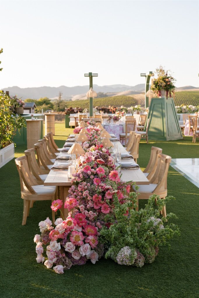 A long outdoor dining table with wooden chairs is decorated with pink and purple flowers, set on green grass with mountains visible in the background.