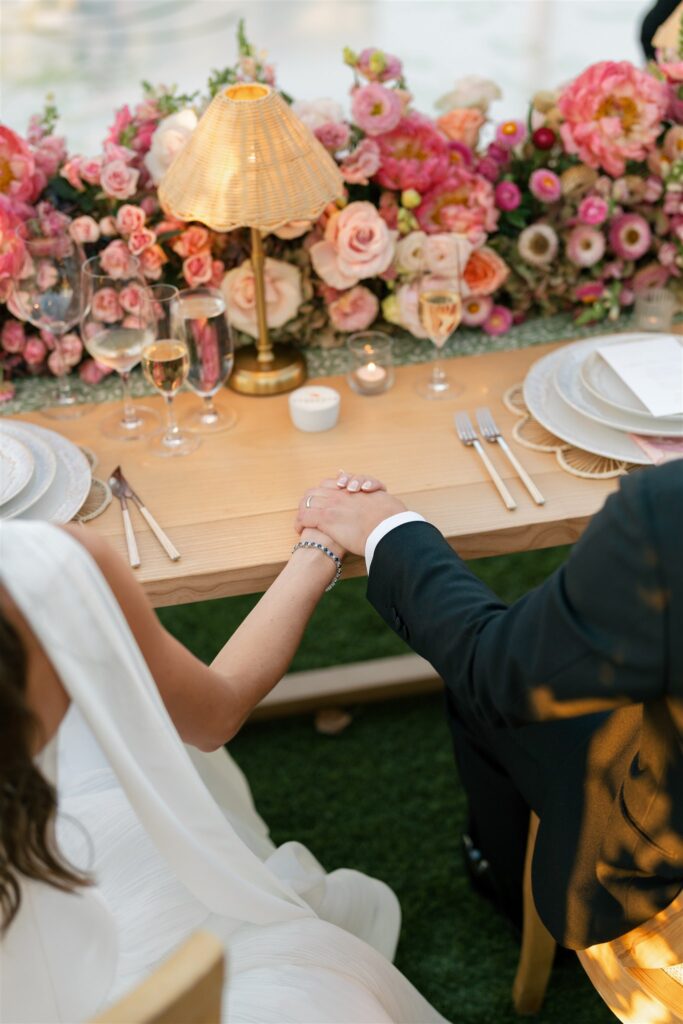 A bride and groom sit at a table decorated with pink flowers, holding hands. Plates, glasses, and a small lamp are on the table.