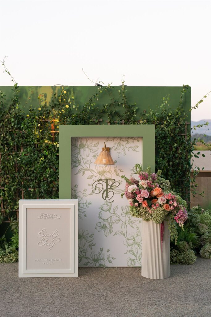 A green-framed floral backdrop with a gold lamp, a monogram, an invitation in a frame, and a tall vase of mixed flowers, set outdoors against a green leafy wall.