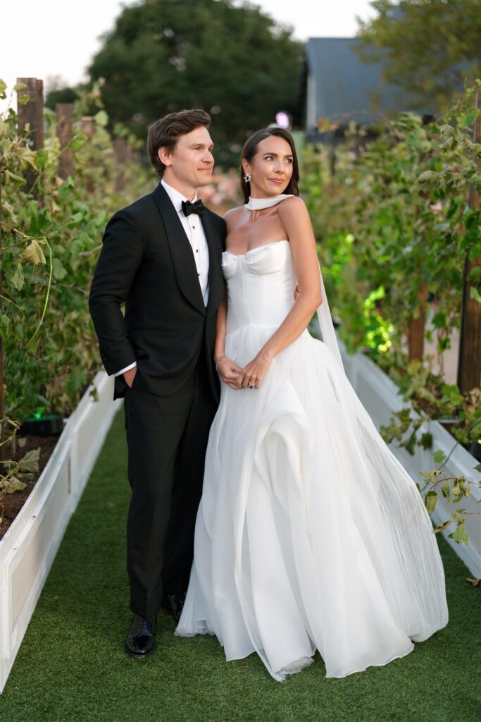 A bride in a white gown and a groom in a black tuxedo stand together outdoors on a grass path lined with green plants.