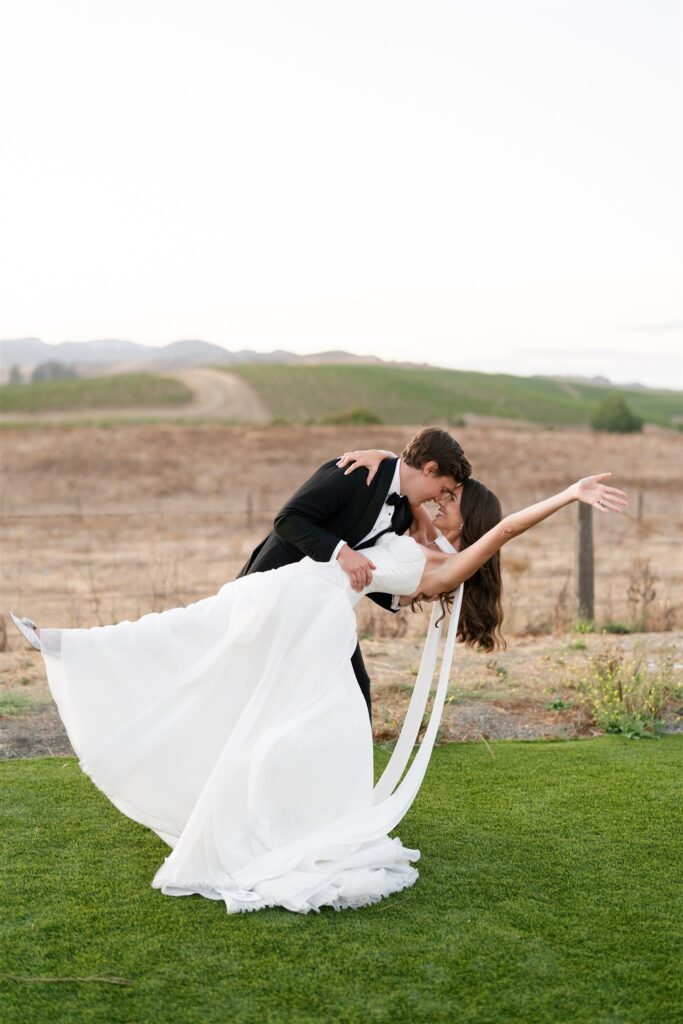 A groom in a black suit dips his bride in a white dress for a kiss outdoors on green grass, with dry fields and hills in the background.