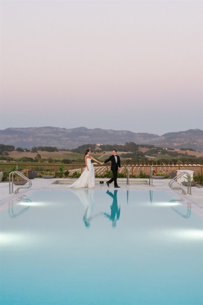 A bride and groom stand hand in hand at the edge of a pool with mountains and vineyards in the background at sunset.