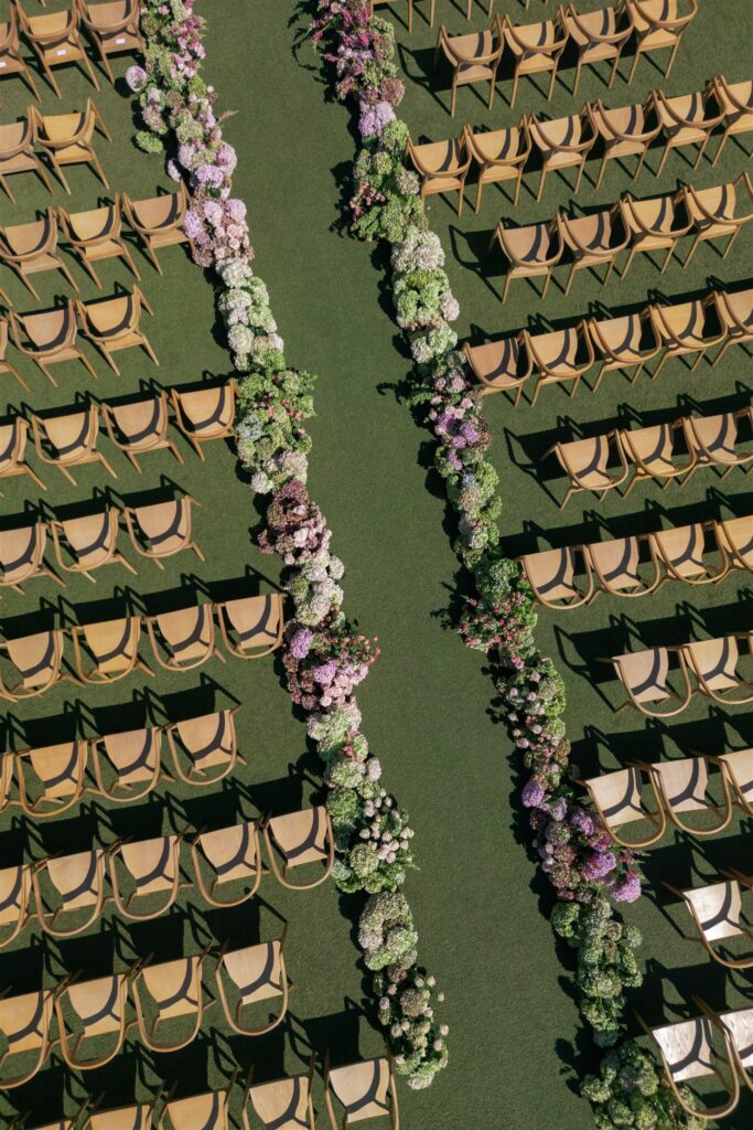 Aerial view of rows of empty wooden chairs arranged beside two parallel aisles lined with green and purple floral arrangements on grass.