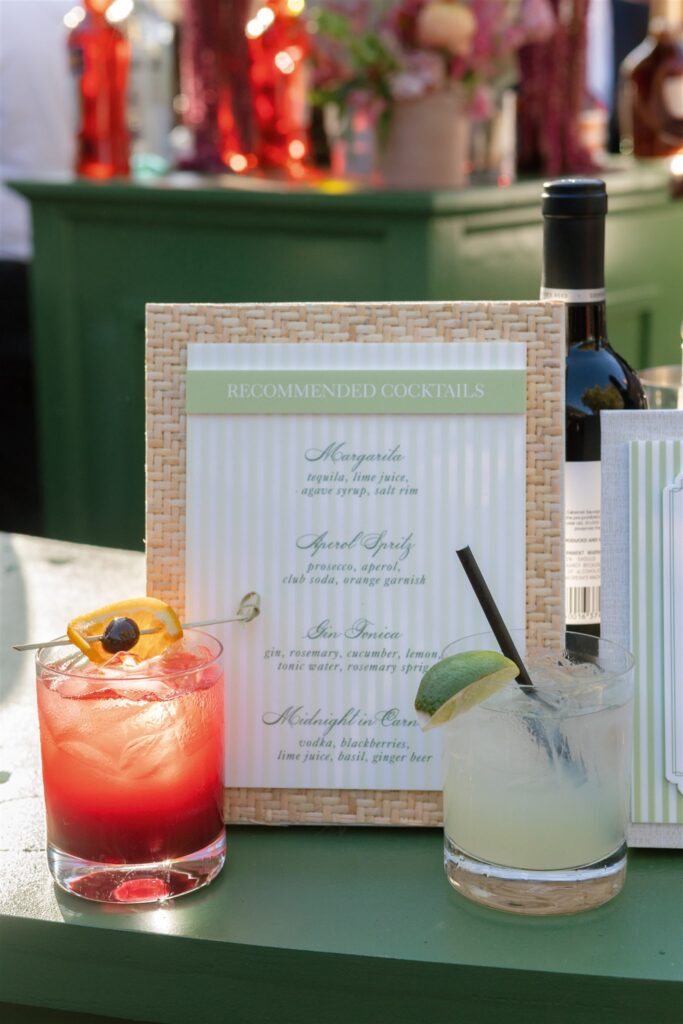 Two cocktails on a green bar next to a sign listing recommended cocktails, with a bottle and blurred background objects visible.